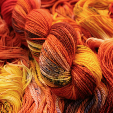 A bright red and orange colored Worsted yarn that has hints of purple and white and some yellow and brown colors. There are more weights of this yarn unwound underneath this skein.