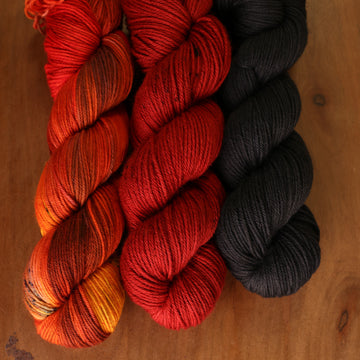 Fire Kit sitting on top of a wooden table, the skeins from left to right are orange with brown streaks, red with brown speckles, and black.