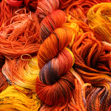 A bright red and orange colored Sport yarn that has hints of purple and white and some yellow and brown colors. There are more weights of this yarn unwound underneath this skein.