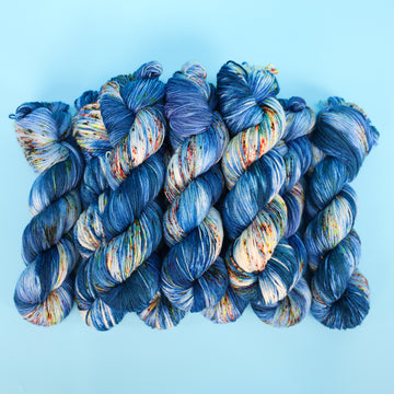 Different weights of a yarn skein colored with different shades of blue and white with multicolored speckles on the portion of the yarn that Is white. all on top of a light blue background.