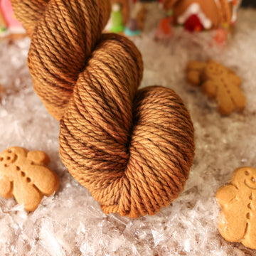 A bulky skein of yarn colored warm and toasty medium brown with very subtle color shifts and speckles. It is sitting on top of fake snow with small gingerbread men and 2 gingerbread houses surround the skein.