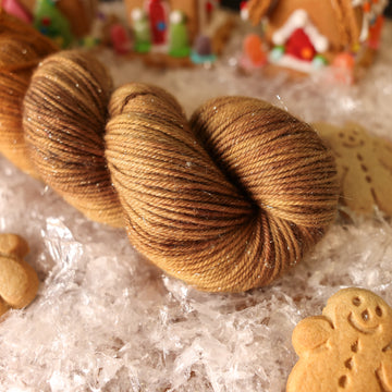 A sparkle DK skein of yarn colored warm and toasty medium brown with very subtle color shifts and speckles. It is sitting on top of fake snow with small gingerbread men and 2 gingerbread houses surround the skein.