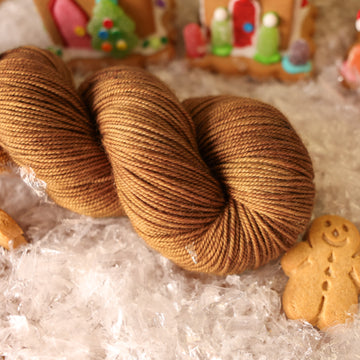 A Sport skein of yarn colored warm and toasty medium brown with very subtle color shifts and speckles. It is sitting on top of fake snow with small gingerbread men and 2 gingerbread houses surround the skein.
