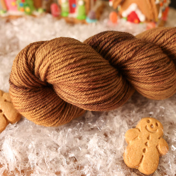 A worsted skein of yarn colored warm and toasty medium brown with very subtle color shifts and speckles. It is sitting on top of fake snow with small gingerbread men and 2 gingerbread houses surround the skein.