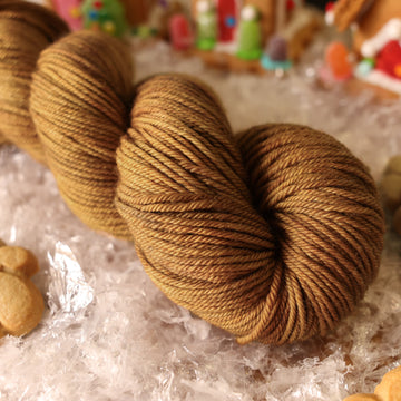 A DK skein of yarn colored warm and toasty medium brown with very subtle color shifts and speckles. It is sitting on top of fake snow with small gingerbread men and 2 gingerbread houses surround the skein.