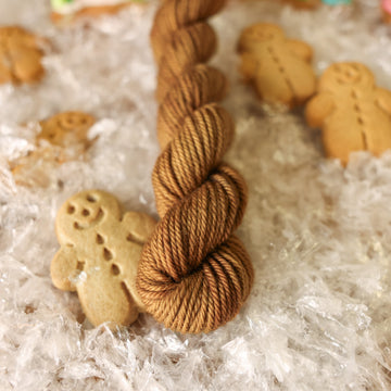 A DK mini skein of yarn colored warm and toasty medium brown with very subtle color shifts and speckles. It is sitting on top of fake snow with small gingerbread men surrounding the skein.