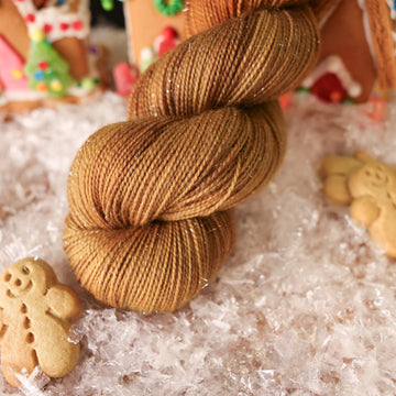 A sparkle fingering skein of yarn colored warm and toasty medium brown with very subtle color shifts and speckles. It is sitting on top of fake snow with small gingerbread men and 2 gingerbread houses surround the skein.