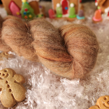 A Suri Alpaca skein of yarn colored warm and toasty medium brown with very subtle color shifts and speckles. It is sitting on top of fake snow with small gingerbread men and 2 gingerbread houses surround the skein.