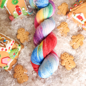 A fingering skein of yarn colored purples, pinks, minty greens, and bright blues, reds, and yellow. It is sitting on top of fake snow with small gingerbread men and three gingerbread houses surround the skein.