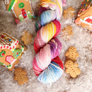 A Bulky skein of yarn colored purples, pinks, minty greens, and bright blues, reds, and yellow. It is sitting on top of fake snow with small gingerbread men and three gingerbread houses surround the skein.