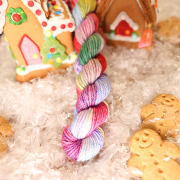 A DK Mini skein of yarn colored purples, pinks, minty greens, and bright blues, reds, and yellow. It is sitting on top of fake snow with small gingerbread men and three gingerbread houses surround the skein.