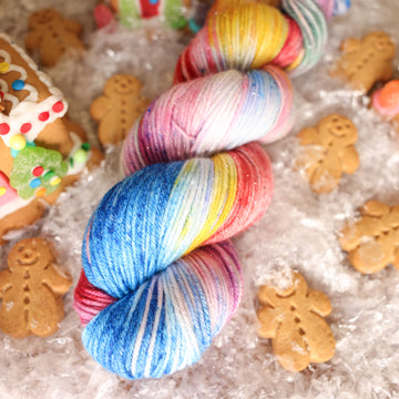 A DK Sparkle skein of yarn colored purples, pinks, minty greens, and bright blues, reds, and yellow. It is sitting on top of fake snow with small gingerbread men and three gingerbread houses surround the skein.