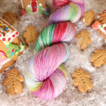 A fingering sparkle skein of yarn colored purples, pinks, minty greens, and bright blues, reds, and yellow. It is sitting on top of fake snow with small gingerbread men and three gingerbread houses surround the skein.