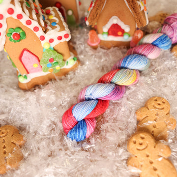 A fingering mini skein of yarn colored purples, pinks, minty greens, and bright blues, reds, and yellow. It is sitting on top of fake snow with small gingerbread men and three gingerbread houses surround the skein.