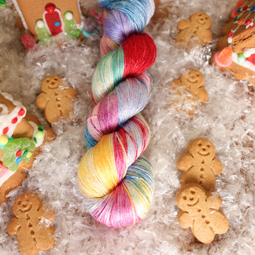 A Silk skein of yarn colored purples, pinks, minty greens, and bright blues, reds, and yellow. It is sitting on top of fake snow with small gingerbread men and three gingerbread houses surround the skein.