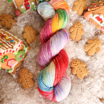 A sport skein of yarn colored purples, pinks, minty greens, and bright blues, reds, and yellow. It is sitting on top of fake snow with small gingerbread men and three gingerbread houses surround the skein.