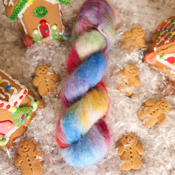 A suri skein of yarn colored purples, pinks, minty greens, and bright blues, reds, and yellow. It is sitting on top of fake snow with small gingerbread men and three gingerbread houses surround the skein.