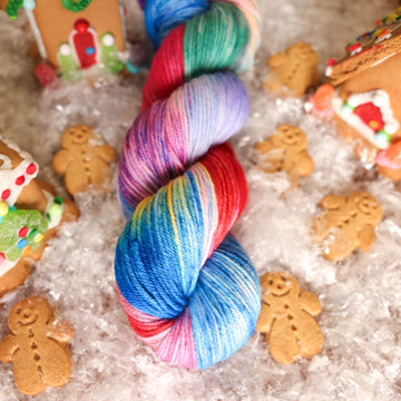 A worsted skein of yarn colored purples, pinks, minty greens, and bright blues, reds, and yellow. It is sitting on top of fake snow with small gingerbread men and three gingerbread houses surround the skein.
