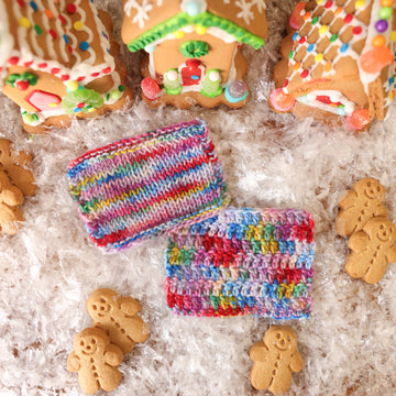 A two knit and crochet swatches colored purples, pinks, minty greens, and bright blues, reds, and yellow. They are sitting on top of fake snow with small gingerbread men and three gingerbread houses surround them.