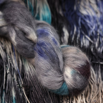 dark grey, light blue, dark blue, with black soft alpaca yarn and speckles of all of those same colors on it, on top of the same yarn but unwound laying flat.