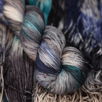 dark grey, light blue, dark blue, with black fingering yarn and speckles of all of those same colors on it, on top of the same yarn but unwound laying flat.
