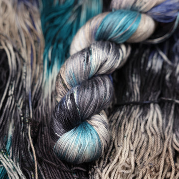 dark grey, light blue, dark blue, with black silk yarn and speckles of all of those same colors on it, on top of the same yarn but unwound laying flat.