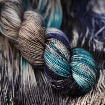dark grey, light blue, dark blue, with black sport yarn and speckles of all of those same colors on it, on top of the same yarn but unwound laying flat.