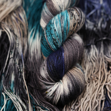 dark grey, light blue, dark blue, with black worsted yarn and speckles of all of those same colors on it, on top of the same yarn but unwound laying flat.