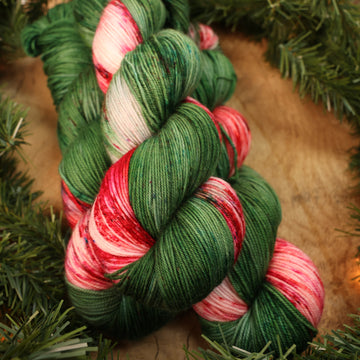 Green and red holiday fingering yarn on a wooden table with Christmas garland surrounding it.