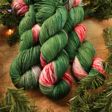 Green and red holiday yarn on a wooden table with Christmas garland surrounding it.