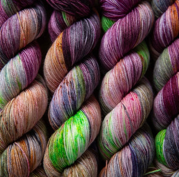 Yarn skeins stacked next to each other that are colored with different shades of darker purples, light oranges, and some lime greens mixed in, along with some other colors.