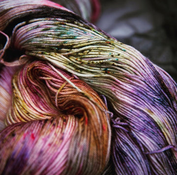 A close up photo of an unwound yarn skein that is colored with different shades of darker purples, light oranges, pinks, dark greens and some other colors mixed in.