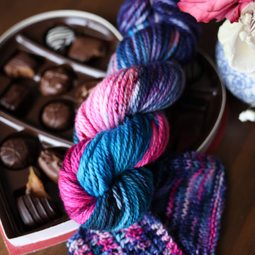 A Bulky skein of yarn colored Bright turquoise and hot pink mix with dark navy and purples with crimson speckles sitting on stop of a box of chocolates. To the side there is a knit and crochet swatches of yarn next to the boxes. In the corner there is a pink rose sitting on a blue and white bag. All is sitting on a wooden table.