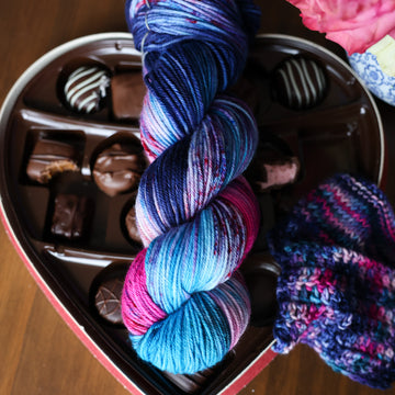A DK skein of yarn colored Bright turquoise and hot pink mix with dark navy and purples with crimson speckles sitting on stop of a box of chocolates. To the side there is a knit and crochet swatches of yarn next to the boxes. In the corner there is a pink rose sitting on a blue and white bag. All is sitting on a wooden table.