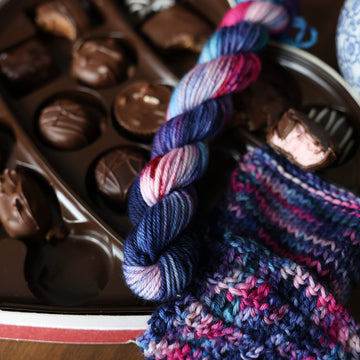 A DK Mini skein of yarn colored Bright turquoise and hot pink mix with dark navy and purples with crimson speckles sitting on stop of a box of chocolates. To the side there is a knit and crochet swatches of yarn next to the boxes. In the corner there is a  blue and white bag. All is sitting on a wooden table.