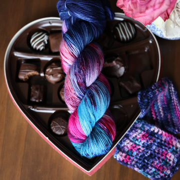 A Fingering skein of yarn colored Bright turquoise and hot pink mix with dark navy and purples with crimson speckles sitting on stop of a box of chocolates. To the side there is a knit and crochet swatches of yarn next to the boxes. All is sitting on a wooden table.
