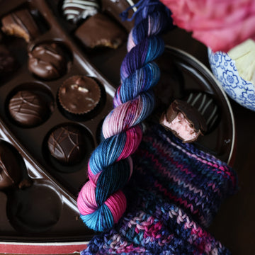 A Fingering Mini skein of yarn colored Bright turquoise and hot pink mix with dark navy and purples with crimson speckles sitting on stop of a box of chocolates. To the side there is a knit and crochet swatches of yarn next to the boxes. In the corner there is a pink rose sitting on a blue and white bag. All is sitting on a wooden table.