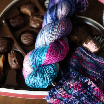 A Silk skein of yarn colored Bright turquoise and hot pink mix with dark navy and purples with crimson speckles sitting on stop of a box of chocolates. To the side there is a knit and crochet swatches of yarn next to the box. All is sitting on a wooden table.