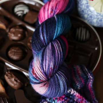 A sparkle DK skein of yarn colored Bright turquoise and hot pink mix with dark navy and purples with crimson speckles sitting on stop of a box of chocolates. To the side there is a knit and crochet swatches of yarn next to the boxes. In the corner there is a blue and white bag. All is sitting on a wooden table.