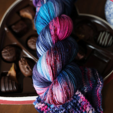 A Sparkle Fingering skein of yarn colored Bright turquoise and hot pink mix with dark navy and purples with crimson speckles sitting on stop of a box of chocolates. To the side there is a knit and crochet swatches of yarn next to the boxes. All is sitting on a wooden table.