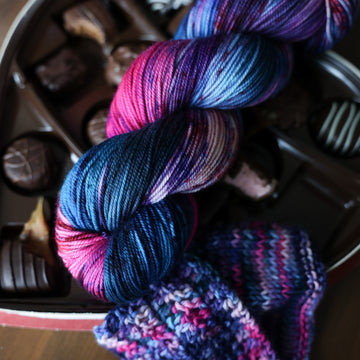 A Sport skein of yarn colored Bright turquoise and hot pink mix with dark navy and purples with crimson speckles sitting on stop of a box of chocolates. Sitting next to the yarn there is a knit and crochet swatches of yarn next to the boxes. All is sitting on a wooden table.