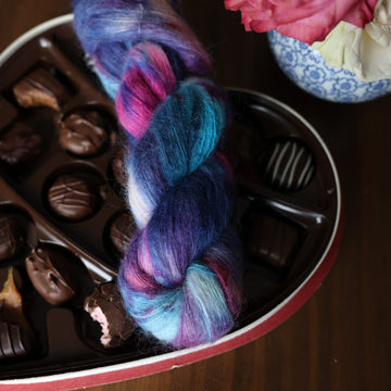 A Suri skein of yarn colored Bright turquoise and hot pink mix with dark navy and purples with crimson speckles sitting on stop of a box of chocolates. In the corner there is a pink rose sitting on a blue and white bag. All is sitting on a wooden table.