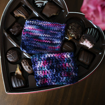 A Knit and crochet swatches of yarn colored Bright turquoise and hot pink mix with dark navy and purples with crimson speckles sitting on top of a box of chocolates. In the corner there is a pink rose, all sitting on top of a wooden table.