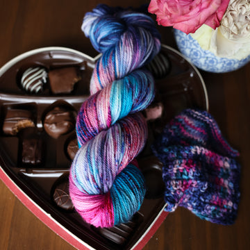 A Worsted skein of yarn colored Bright turquoise and hot pink mix with dark navy and purples with crimson speckles sitting on stop of a box of chocolates. To the side there is a knit and crochet swatches of yarn next to the boxes. In the corner there is a pink rose sitting on a blue and white bag. All is sitting on a wooden table.