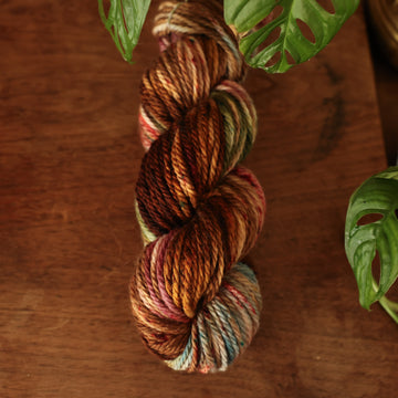 Bulky yarn colored with different shades of dark and light brown with little hints of red and green, on top of a wooden table with some of a plant peeking into the frame.