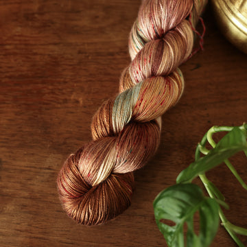 Silk yarn colored with different shades of dark and light brown with little hints of red and green, on top of a wooden table with some of a plant peeking into the frame.