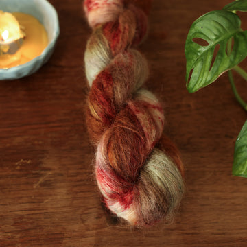 Suri alpaca yarn colored with different shades of dark and light brown with little hints of red and green, on top of a wooden table with some of a plant peeking into the frame.