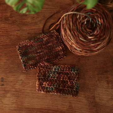 Crochet and Knit swatches colored with different shades of dark and light brown with little hints of red and green, on top of a wooden table with some of a plant peeking into the frame, and a caked version of this yarn in the corner.