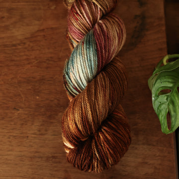 Worsted yarn colored with different shades of dark and light brown with little hints of red and green, on top of a wooden table with some of a plant peeking into the frame.