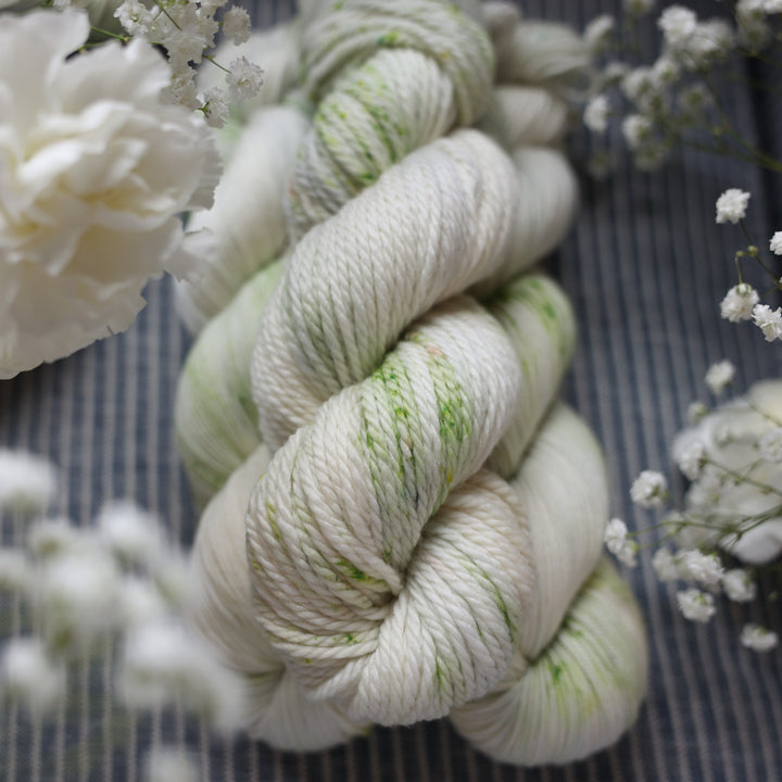 Bulky yarn colored a white and very light green with some darker green speckles. These skeins are on top of a faded navy blue and white striped fabric with white flowers like roses and babies breath around the yarn.